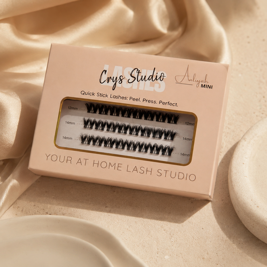 A boxed pair of black false eyelashes on a blush pink package labeled 'Crys Studio' and 'Aaliyah Mini,' styled on draped champagne satin fabric with a round beige dish nearby.